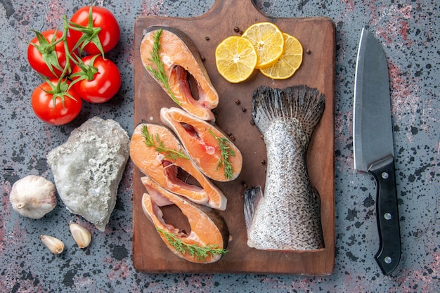 Close up view of raw fishes lemon slices greens pepper on wooden cutting board and foods knife on blue black colors table