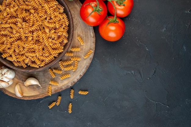 Close up view of raw pastas inside and outside brown pot garlics tomatoes on the right side on dark blue color background