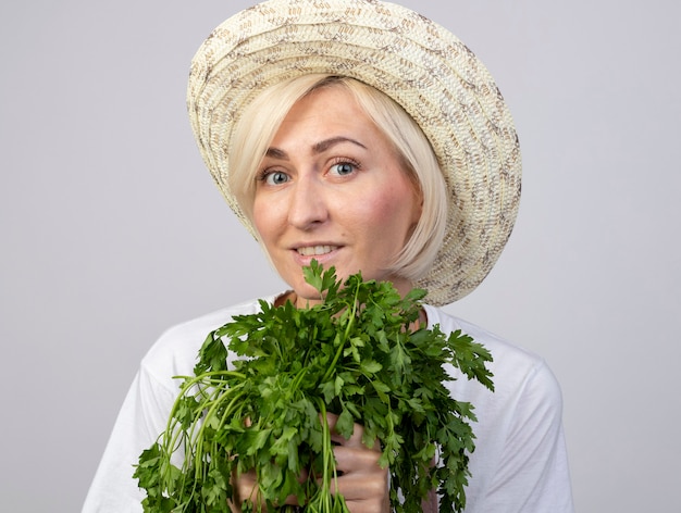 Close-up view of smiling middle-aged blonde gardener woman in uniform wearing hat holding two bunches of coriander 