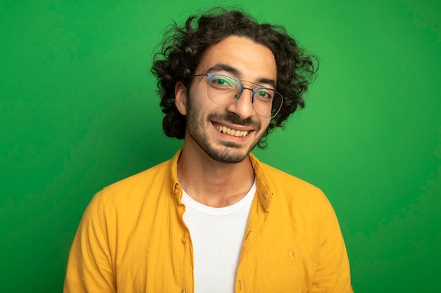 Close-up view of smiling young handsome man wearing glasses looking at front isolated on green wall