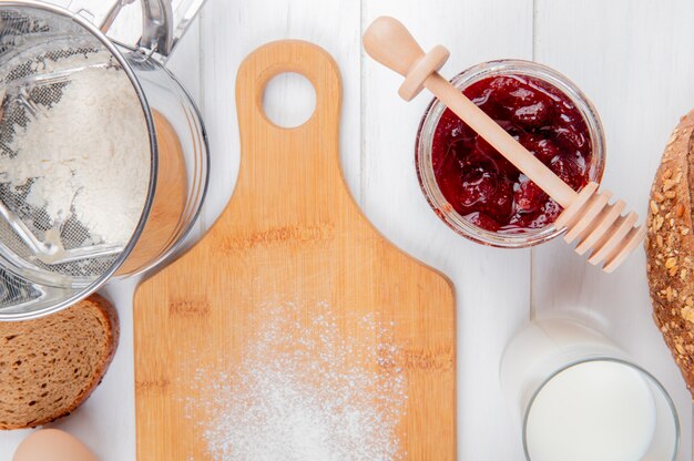 Close-up view of strawberry jam in jar flour slice of rye bread glass of milk and cutting board on wooden surface