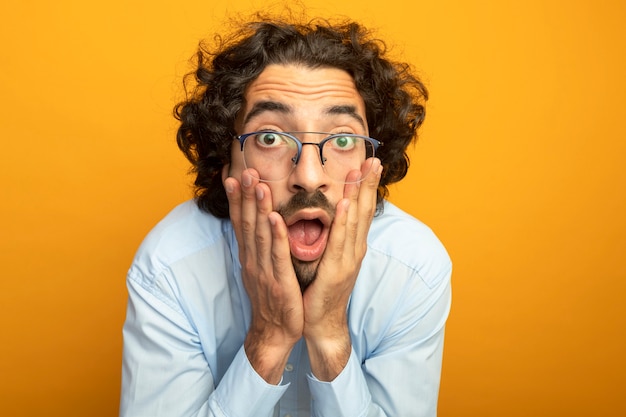 Close-up view of surprised young handsome man wearing glasses putting hands on face looking at front isolated on orange wall