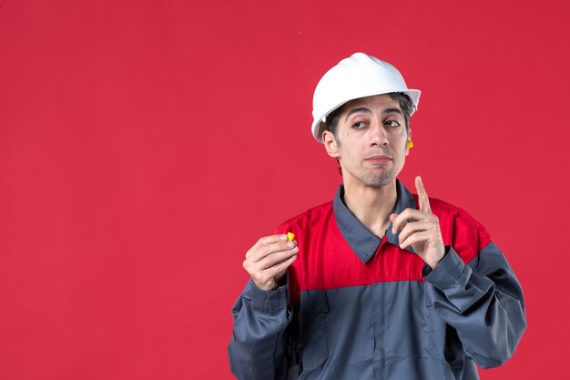 Close up view of thinking young worker in uniform with hard hat and holding earplugs on isolated red wall