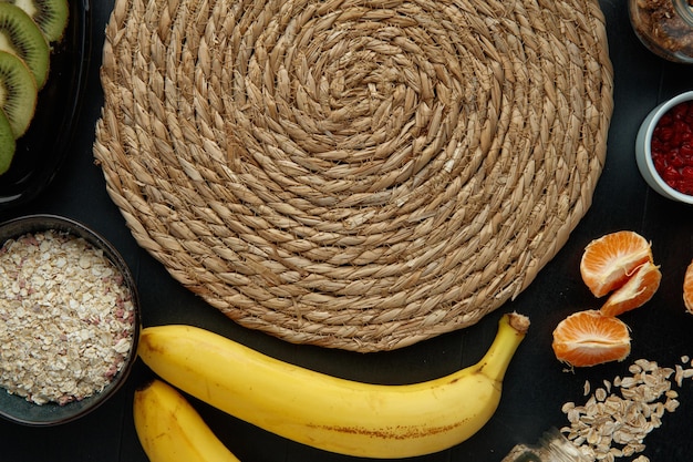 Close up view of trivet with citrus fruits as banana kiwi tangerine oat seeds walnut red currant on black background