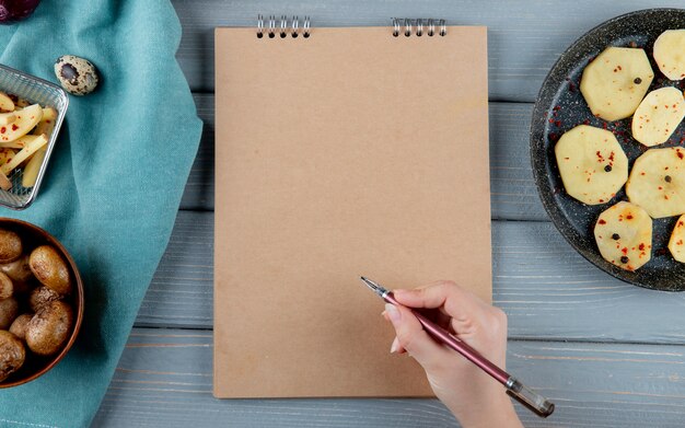 Close up view of woman hand holding pen and note pad with baked and sliced potatoes around on wooden background with copy space