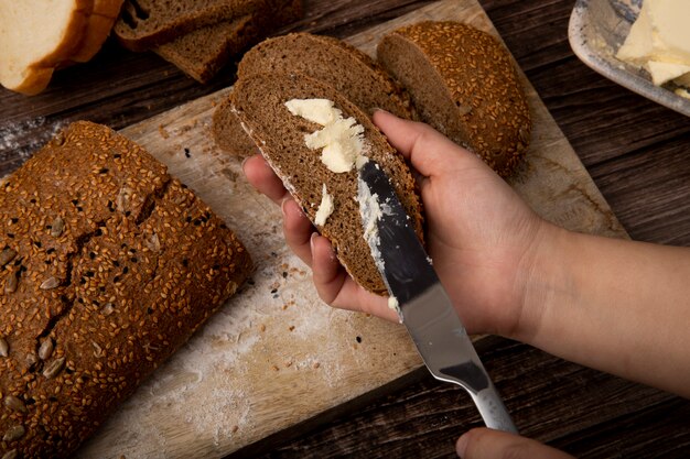 Close-up view of woman hans spreading butter on bread with knife and cut bread on wooden surface and background