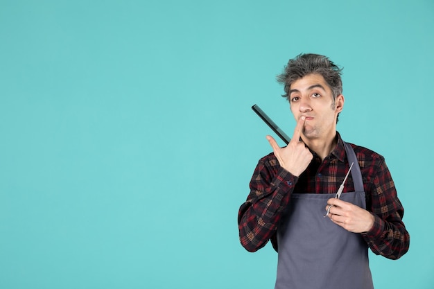 Close up view of young thinking male barber wearing gray apron and holding comb scissor on blue color surface