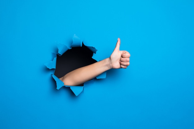 Close up of woman hand with thumbs up gesture through blue hole in paper wall.