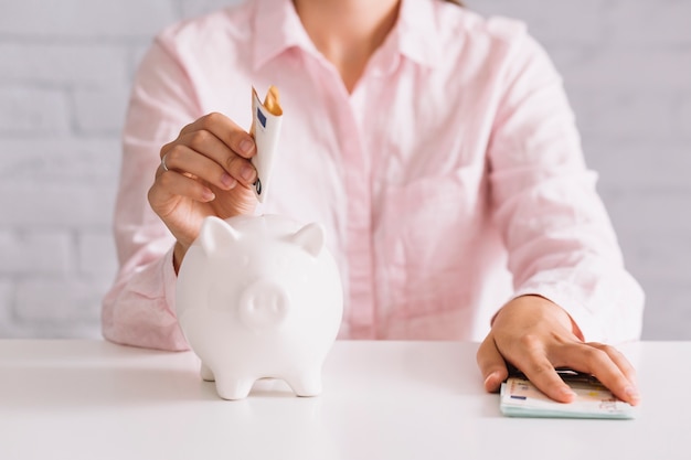 Close-up of woman inserting euro note in white piggybank