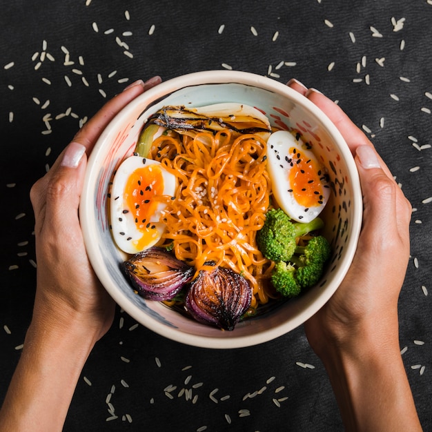 Free photo close-up of woman's hand holding bowl of noodles with eggs; onion; broccoli in bowl on black background