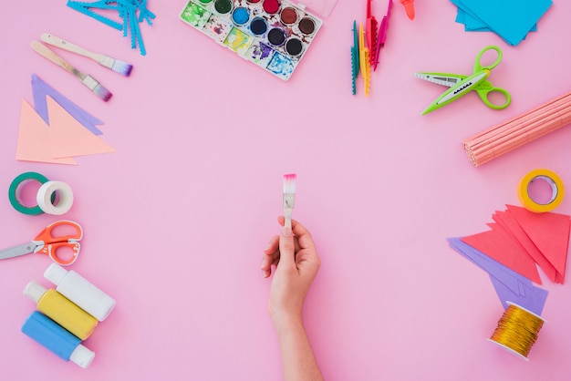 Free photo close-up of woman's hand holding paint brush with water color palette; paintbrush; paper; scissor on pink backdrop