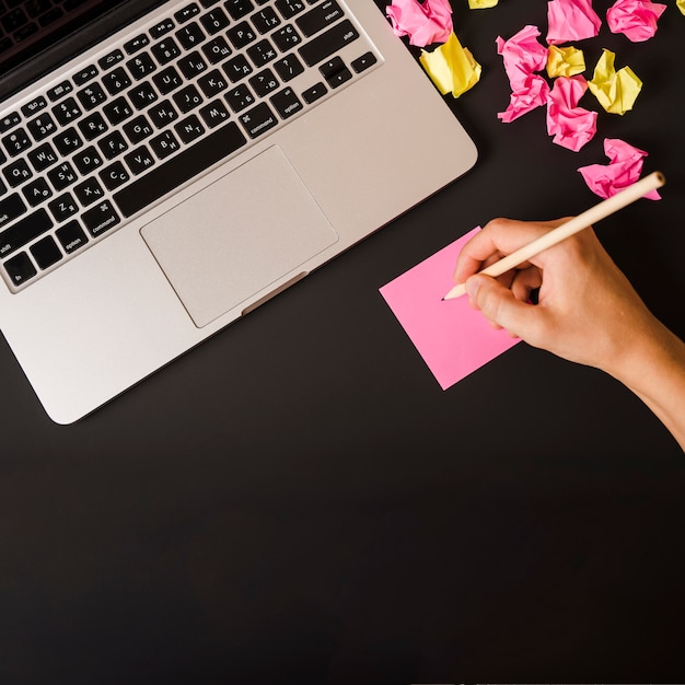 Free photo close-up of woman's hand writing on adhesive note with laptop and crumpled paper on black background