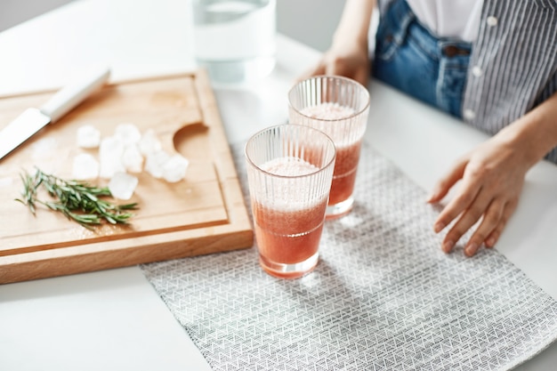 Close up of woman's hands glasses with grapefruit detox diet smoothie rosemary and ice pieces on wooden desk.