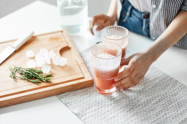 Close up of woman's hands glasses with grapefruit detox diet smoothie rosemary and ice pieces on wooden desk.