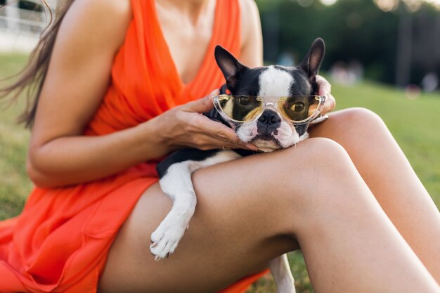 Close up of woman sitting on grass in summer park holding boston terrier, funny dog wearing stylish sunglasses, girl playing with pet, having fun