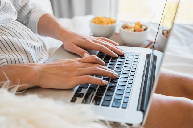 Close-up of woman typing on laptop with breakfast on bed