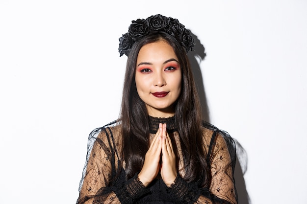 Close-up of young asian woman in black gothic dress and wreath holding hands in pray, girl wearing witch costume and celebrating halloween.