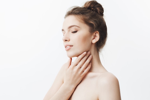 Free photo close up of young attractive caucasian woman with dark hair in stylish hairstyle and naked shoulders touching face with fingers, gently smiling, posing with relaxed face expression and closed eyes.