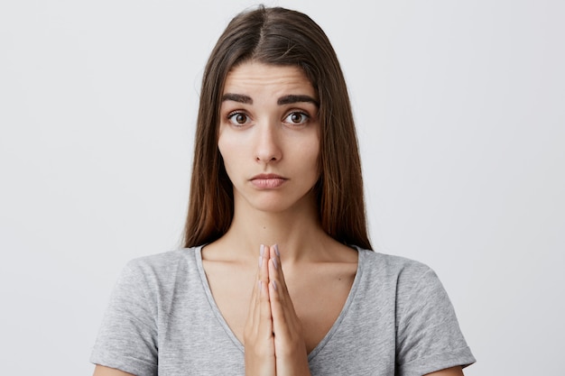 Free Photo close up of young attractive sexy caucasian woman with long dark hair in casual gray t-shirt holding palms together with pleasing expression,