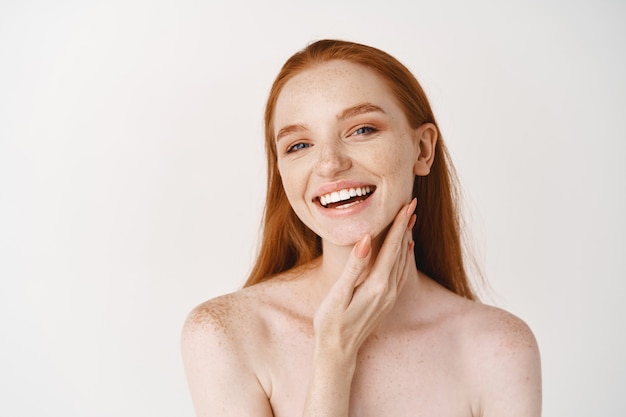 Close-up of young beautiful redhead woman smiling at at front, touching perfect clean skin on face and looking happy, standing naked over white wall