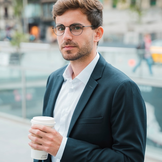 Close-up of a young businessman holding disposable coffee cup in hand looking at camera