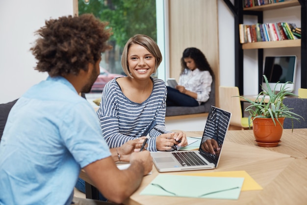 Free photo close up of young cheerful student girl with light hair in bob hairstyle sitting on meeting with friend from university, doing team project, searching for information on laptop.