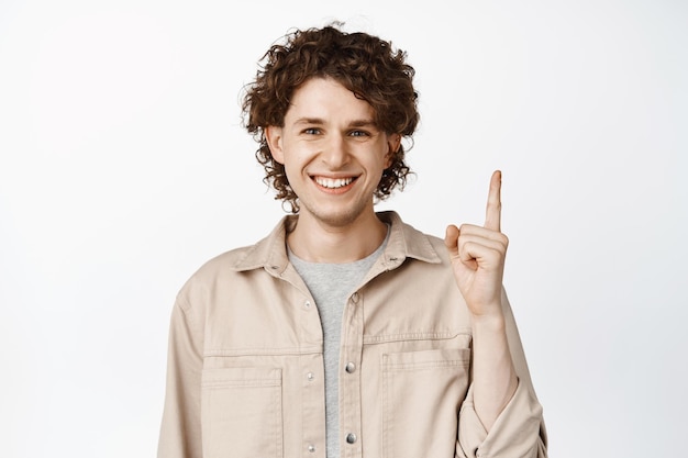 Free photo close up of young happy guy showing advertisement pointing finger up standing over white background