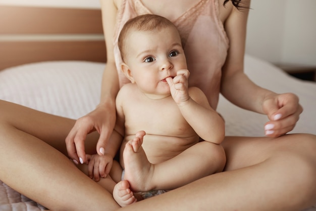 Close up of young mother in sleepwear and her newborn baby sitting on bed in morning smiling playing together