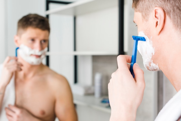 Close-up of a young shirtless man shaving with blue razor in front of mirror