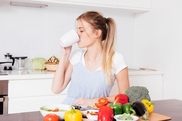 Close-up of a young woman drinking the coffee with fresh colorful vegetables on wooden table