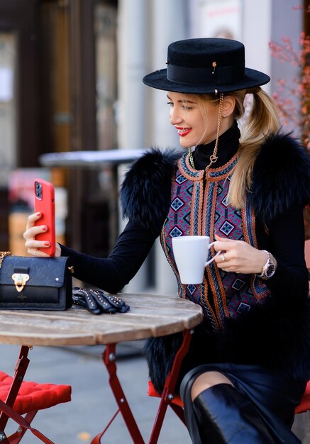 Close view of attractive woman in black hat with chain dressed in trendy vest with fur and embroidery sitting at table using mobile phone and taking selfie while drinking coffee in cafeteria