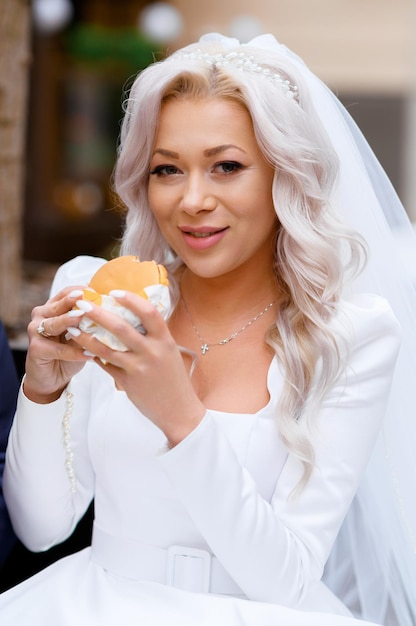 Close view of cheerful and pretty bride with hairstyle and accessories on head wearing in design wedding dress with long sleeves and belt on waist holding sandwich while looking at camera outdoor