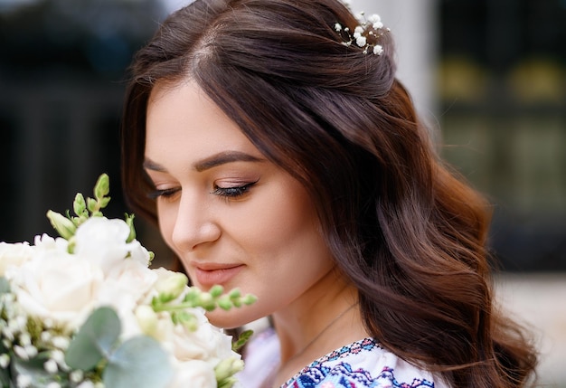 Close view of lovely face of young girl which have brunette curly hair and cute makeup holding bridal flowers and looking down while smelling it