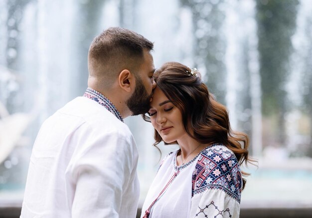 Close view of stunning and pensive woman with natural makeup and curly hair dressed in shirt with colorful pattern bending head forward while man kissing her face on background of fountains