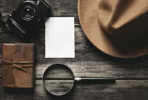 Free photo closed notebook in leather cover, sheet of white paper, felt brown hat, camera and big magnifier isolated on black aged wood table