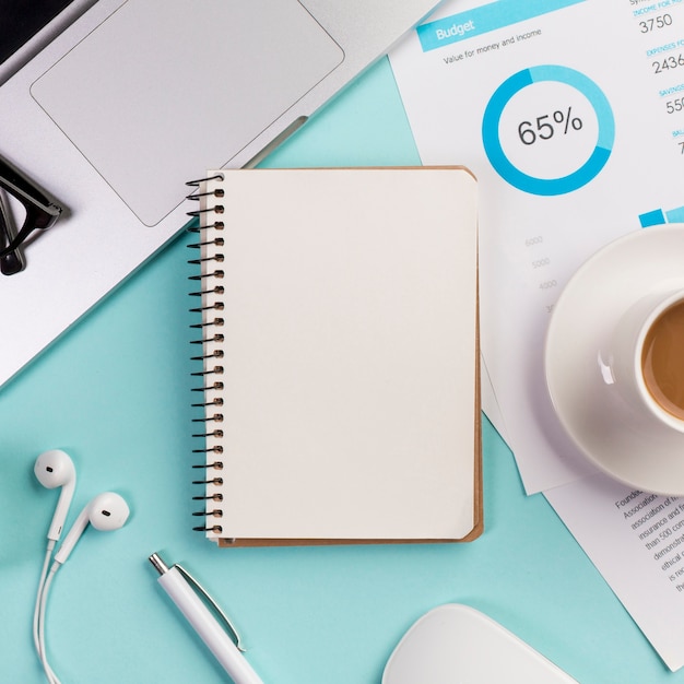 Closed spiral notepad with laptop,earphone,pen,mouse and coffee cup on budget plan on blue desk