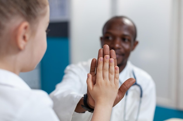 Closeup of african american pediatrician doctor giving high five to young girl during clinical appointment. Therapist man explaining healthcare treatment working in hospital office. Medical service