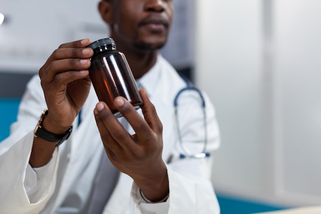 Closeup of african american pediatrician doctor holding pills bottle in hand during clinical appointment in hospital office. Therapist man discussing medication treatment. Healthcare service