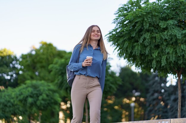 Closeup attractive woman in motion with takeaway coffee on city street. Portrait blonde girl holding paper cup with hot drink outdoor.