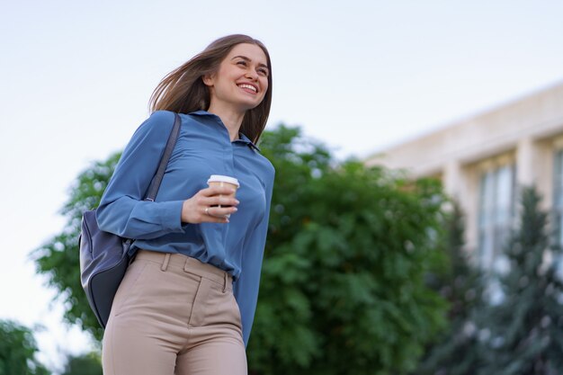 Closeup attractive woman in motion with takeaway coffee on city street. Portrait blonde girl holding paper cup with hot drink outdoor.