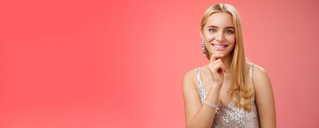 Free photo closeup charming tender young cute blond girl in silver stylish party dress touching chin smiling friendly listen curiously interesting conversation standing red background amused entertained