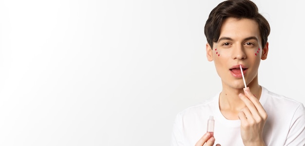 Closeup of gender fluid person with glitter on face applying lip gloss lipstick and looking at camera standing against white background