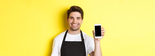 Free photo closeup of handsome waiter in black apron showing smartphone screen recommending app standing over y