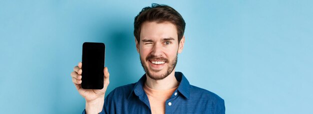 Closeup of handsome young man smiling winking and showing empty mobile phone screen standing in casu