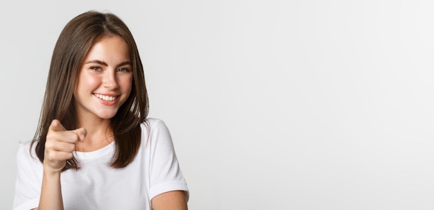 Free photo closeup of happy smiling brunette girl pointing at camera standing white background