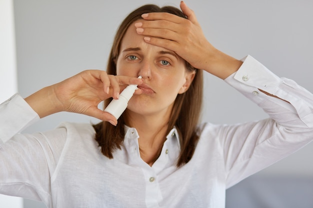 Free Photo closeup of ill young adult woman using nasal spray, suffering from runny nose and terrible headache, looking at camera, touching her forehead, having flu and high temperature.