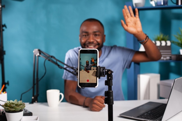 Closeup of live vlog setup with smarthone on stand filming content creator waving hand to followers in online morning show. Selective focus on vlogger saying hello at audience sitting at desk.