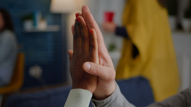 Closeup of multiethnic friends giving high five during celebration party while sitting on sofa late ...