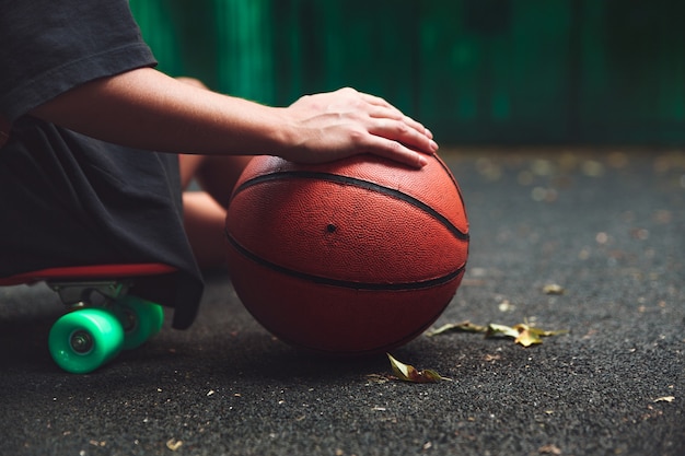 Closeup photo basketball ball with girl sitting on plastic orange penny shortboard on asphalt