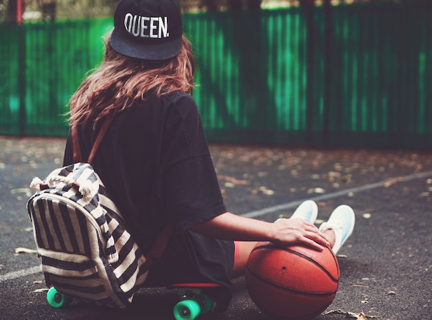 Closeup photo basketball ball with girl sitting on plastic orange penny shortboard on asphalt